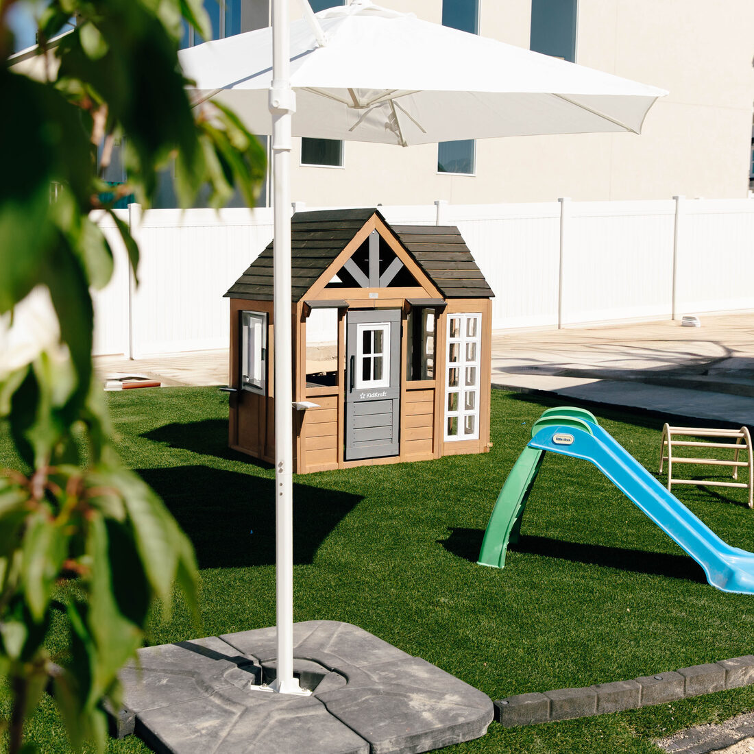 The outdoor play area on turf: a wooden playhouse, a slide, and a parasol for shade.