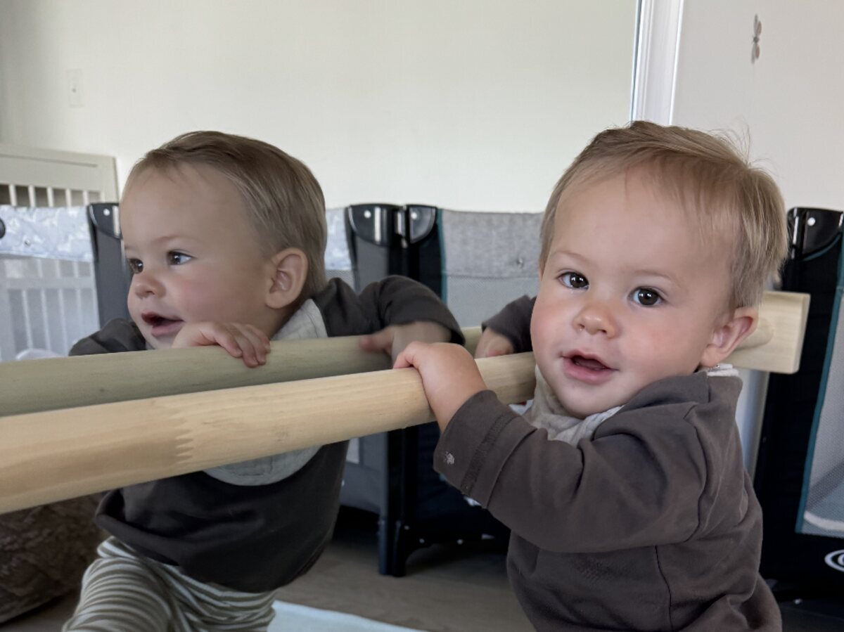 An infant standing at a low wooden barre, gripping it with both hands, reflected in the mirror behind.