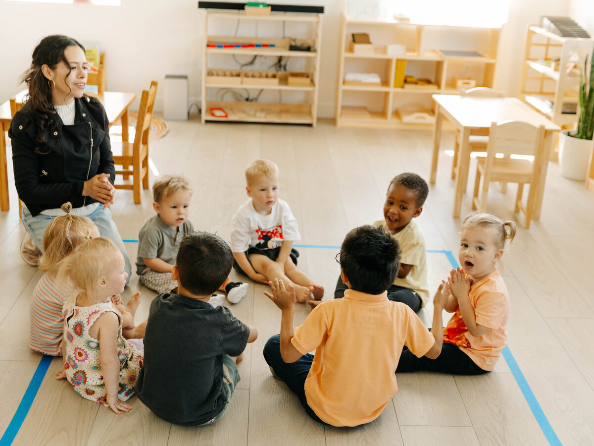 A teacher sitting on the floor with eight children gathered in a circle around her.