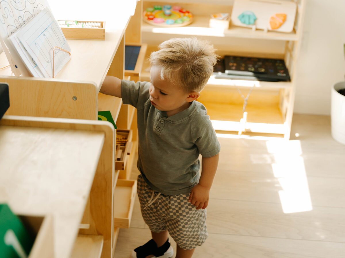 A toddler at an open shelf in the afternoon light, reaching for a tray of materials.